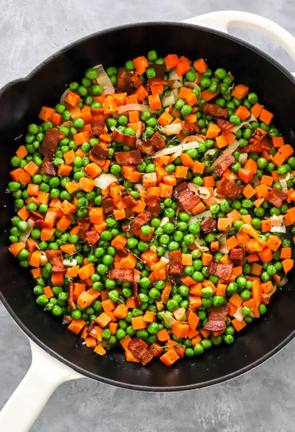 Cooked peas and carrots with bacon in a white skillet.