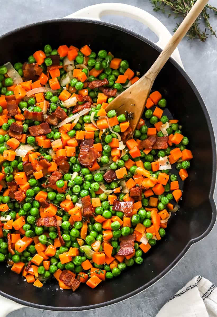 Chopped peas and carrots with crispy pieces of bacon in it in a skillet with a wooden spoon in the pan and some thyme sprigs behind it.