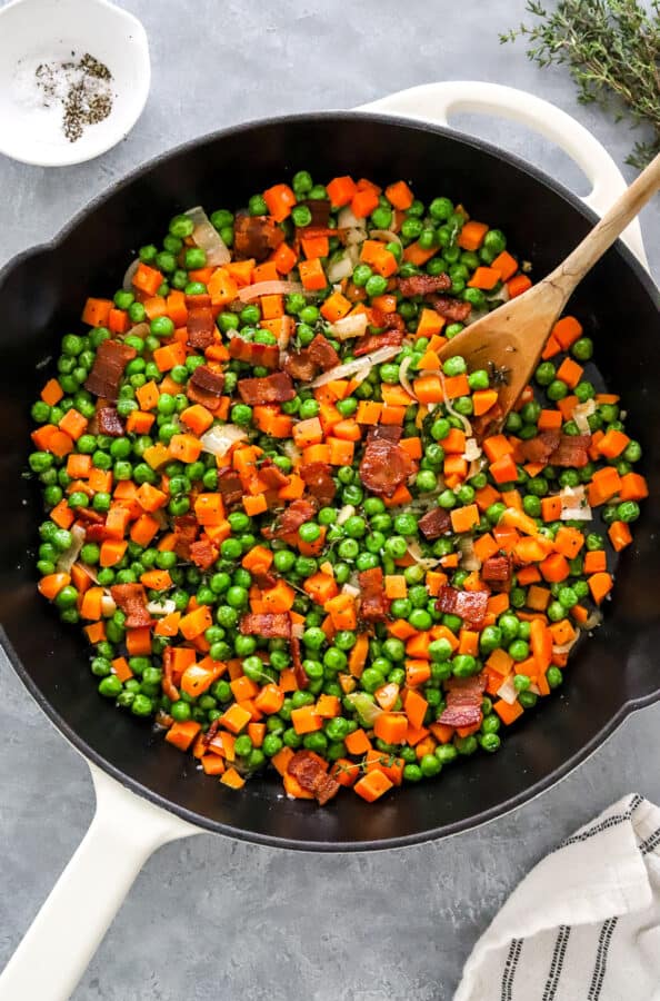 Peas and carrots in a white cast iron pan with bacon with salt and pepper in a bowl and fresh thyme behind it.