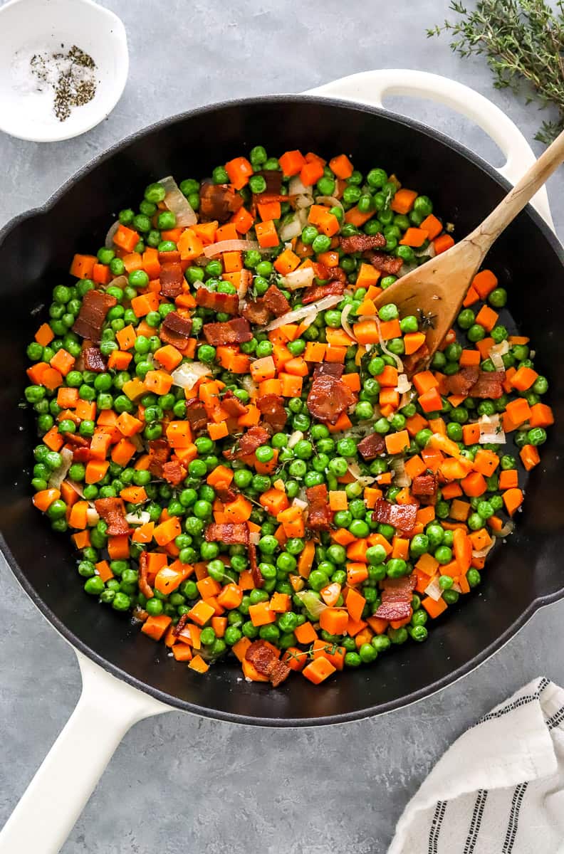 Buttered carrots with peas in a white cast iron pan with bacon with salt and pepper in a bowl and fresh thyme behind it.
