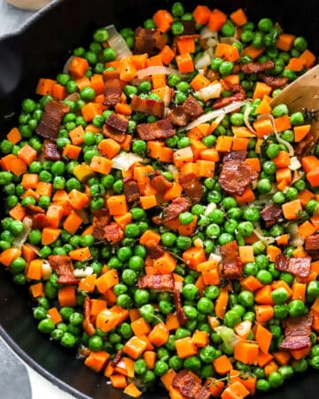 Butteres carrots peas and bacon with onion cooked in a skillet with a wood spoon in the pan and salt and pepper in a wood bowl behind it.