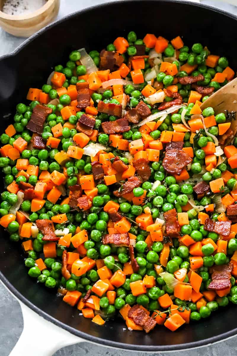 Buttered carrots peas and bacon with onion cooked in a skillet with a wood spoon in the pan and salt and pepper in a wood bowl behind it.