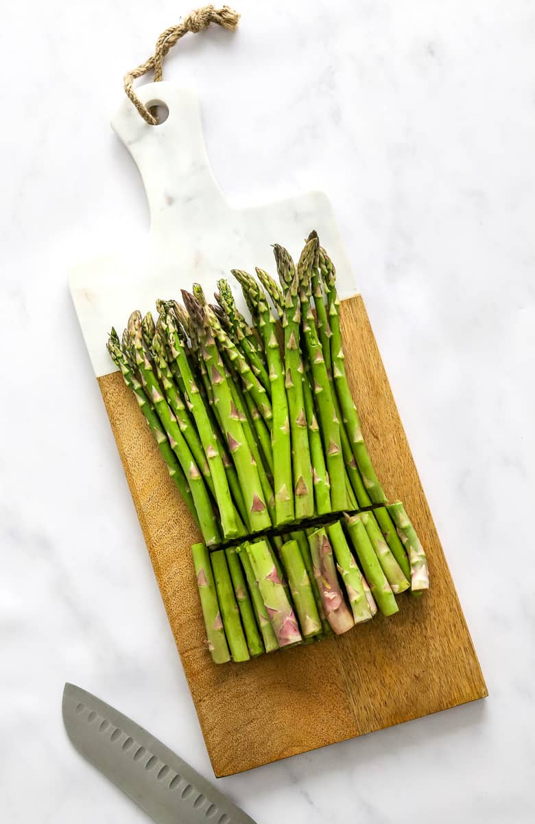 Fresh uncooked asparagus spears on a long cutting board with the ends cut off but still on the board in front of the asparagus. 