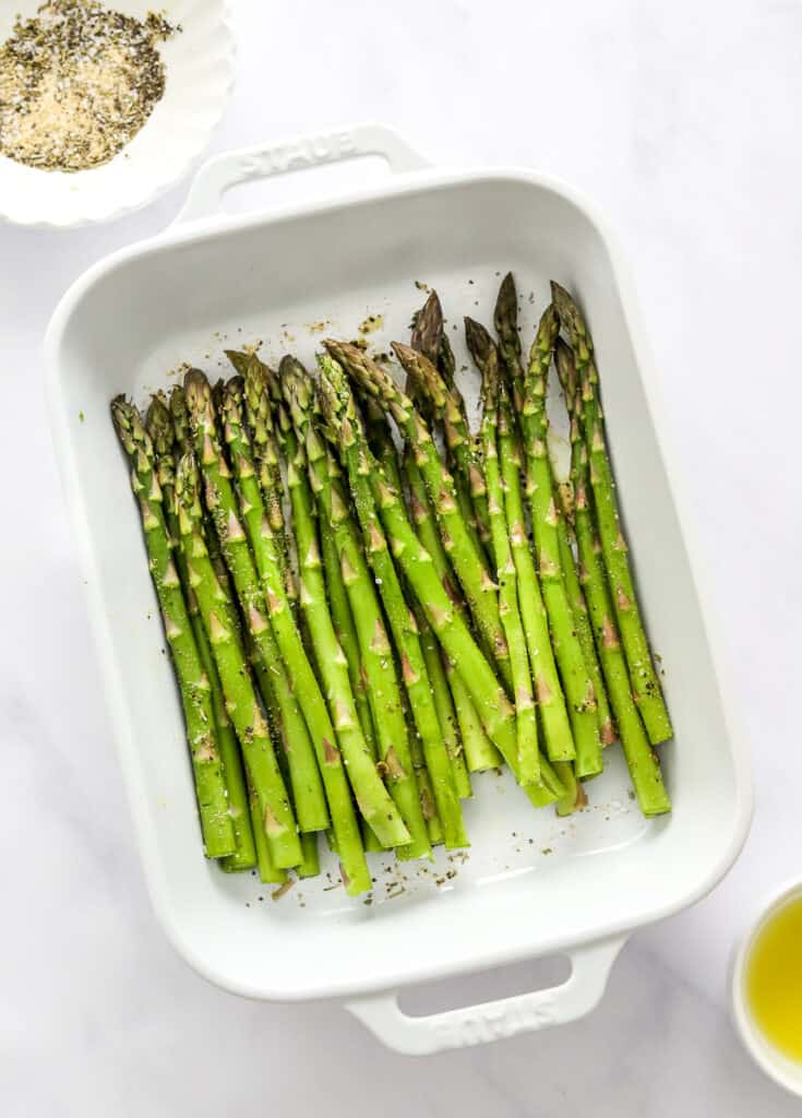 Uncooked, seasoned asparagus in a rectangle white dish with a bowl of seasoning behind it. 