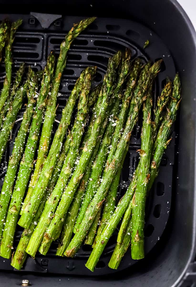Long green air fried asparagus in an air fryer basket with a sprinkle of grated cheese on it. 