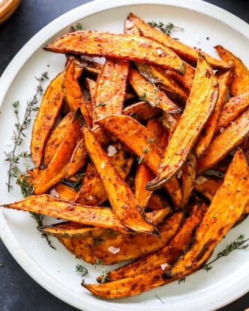 Sweet potato wedges on a grey plate with fresh thyme sprigs on the plate and dipping sauces in bowls around them.