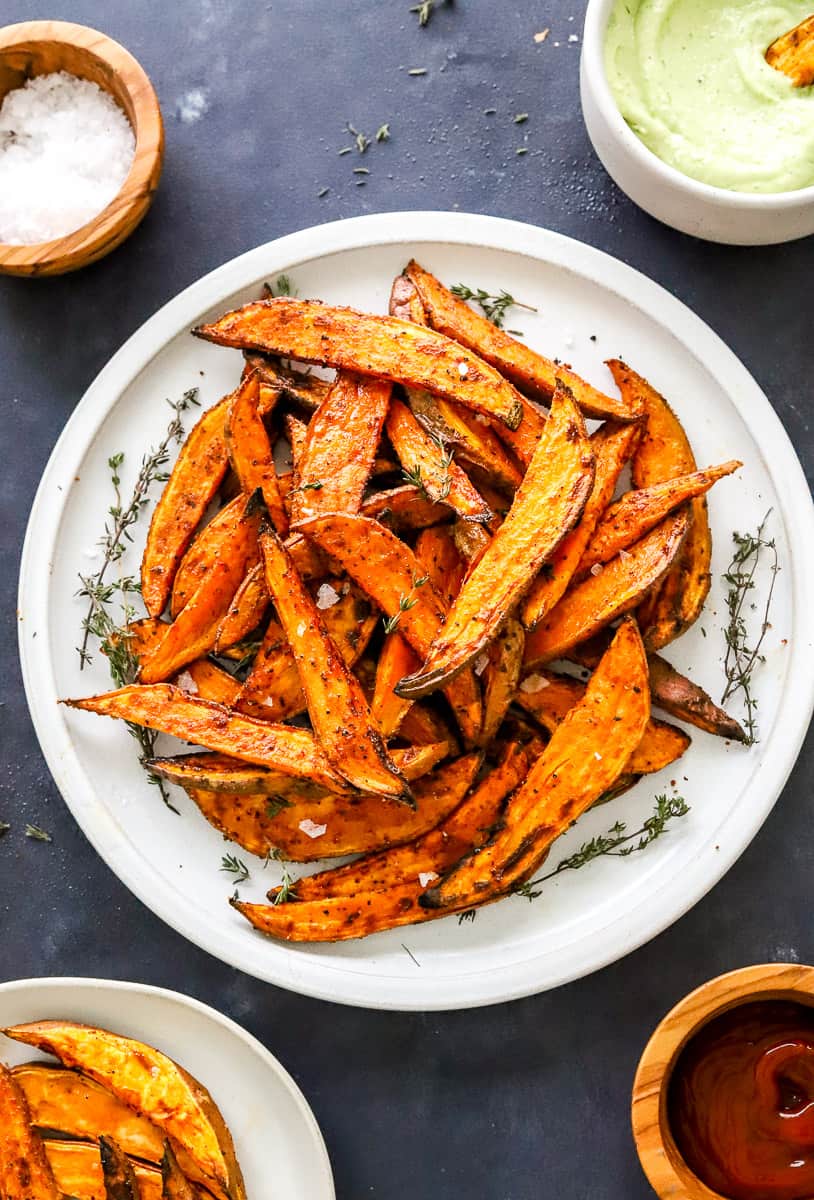 Plate full of air fryer sweet potato wedges with a dipping sauce and salt in bowls behind it and ketchup and more fries on a plate in front of it.
