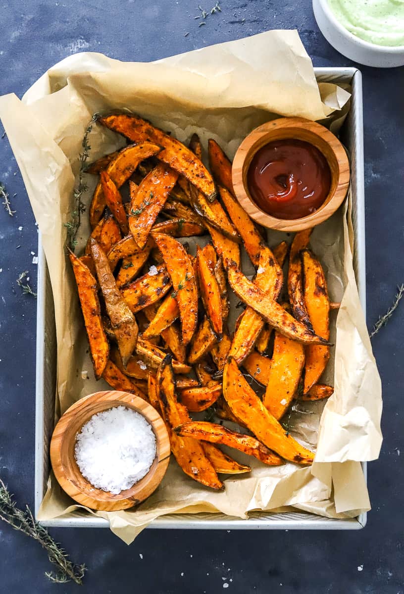 Air fried sweet potato wedges on top of brown paper in a rectangle tray with ketchup in a bowl and salt in a bowl next to it on the tray. 