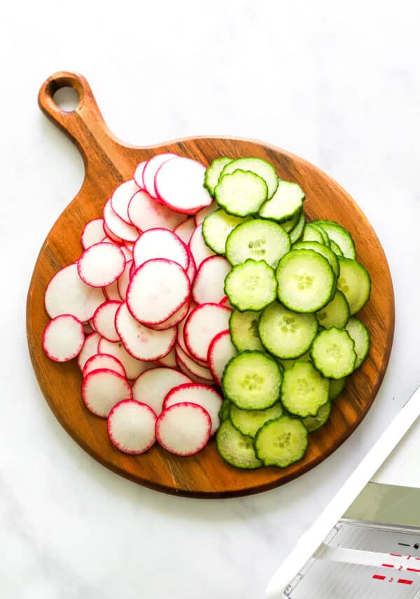Round brown cutting board with a pile of sliced radishes and cucumbers on it with a white Mandolin in front of it.