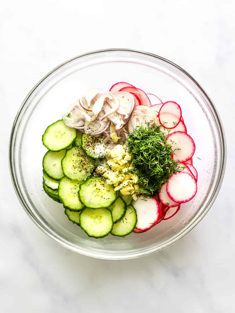 Sliced radishes, sliced cucumber, sliced shallots, chopped garlic and dill in a round glass mixing bowl.