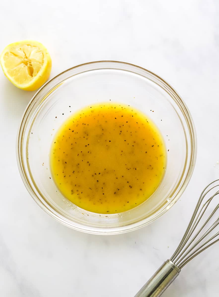 Round glass bowl of dijon lemon dressing with a whisk in front of it and a squeezed half of lemon behind it.