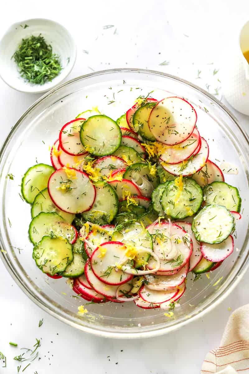 Cucumber radish salad with dill and lemon zest sprinkled on it with a bowl of chopped dil and more dressing behind it.