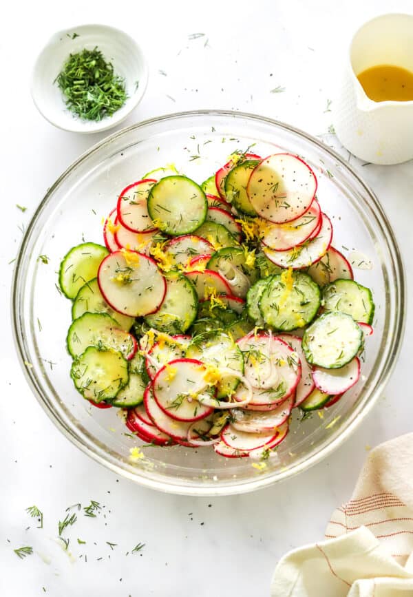 Glass serving bowl filled with sliced radishes and cucumber with lemon zest and dill sprinkled over it with more chopped dill and dressing behind it and a stripped linen in front of it.