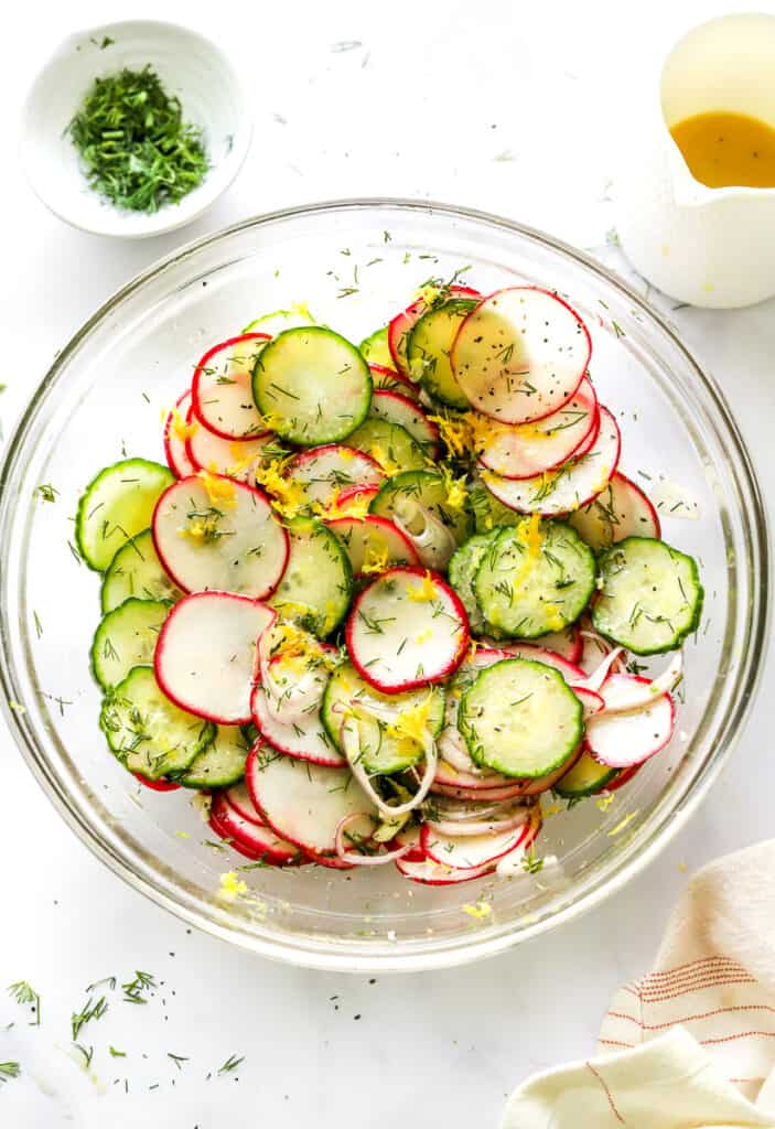 Sliced radish and cucumbers with lemon, dill, and shallots in a glass bowl with a bowl of dill and jar of dressing behind it.