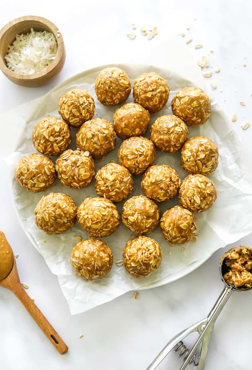Rolled peanut butter oatmeal balls on parchment paper on a plate with a scoop with more of the mixture a spoon with peanut butter in front of it and a bowl of shredded coconut behind it.