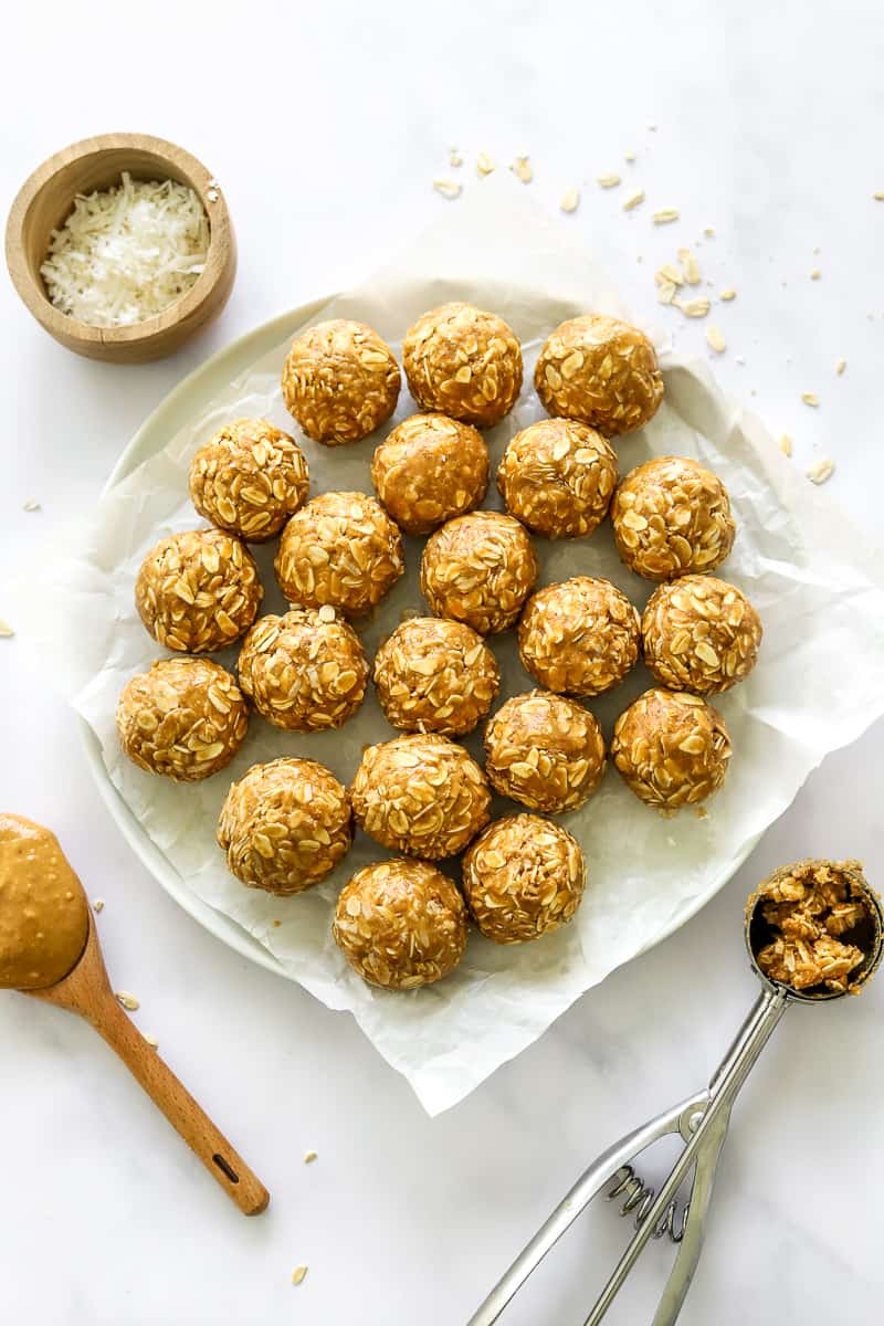 Plate full of peanut butter oatmeal energy bites with a cookie scoop with more of the mixture in it and a wood spoon full of peanut butter infant of it and more coconut behind it.