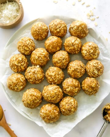 Peanut butter energy bites with oats on a parchment lined plate with a scoop of it and a wood spoon infront of it and a brown bowl of coconut shreds behind it.