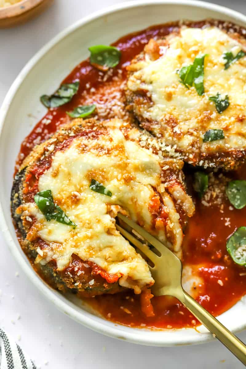Large air fryer eggplant slices on a plate on top of red sauce with cheese and basil with a fork cutting through on of the slices of eggplant. 