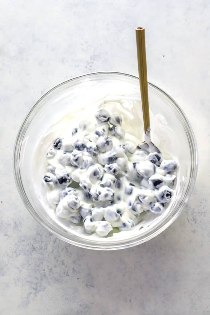 Blueberries covered in yogurt in a glass bowl with a gold spoon in it. 