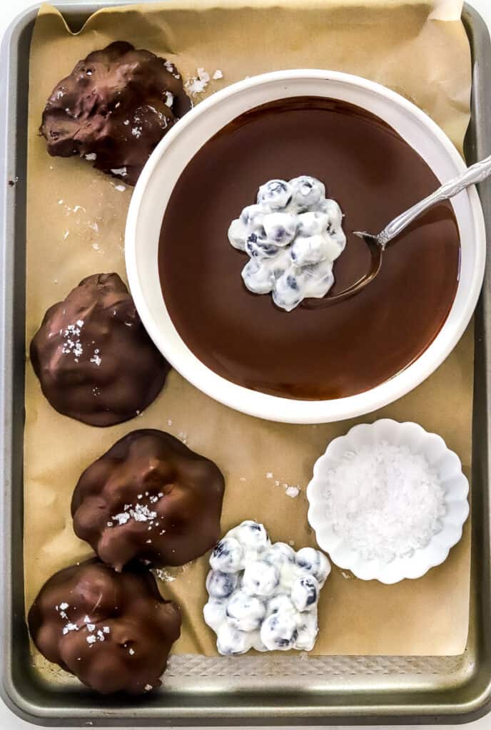 Sheet pan lined with parchment paper with a bowl of melted chocolate with a yogurt blueberry clusters in it with more chocolate clusters on the pan around it. 