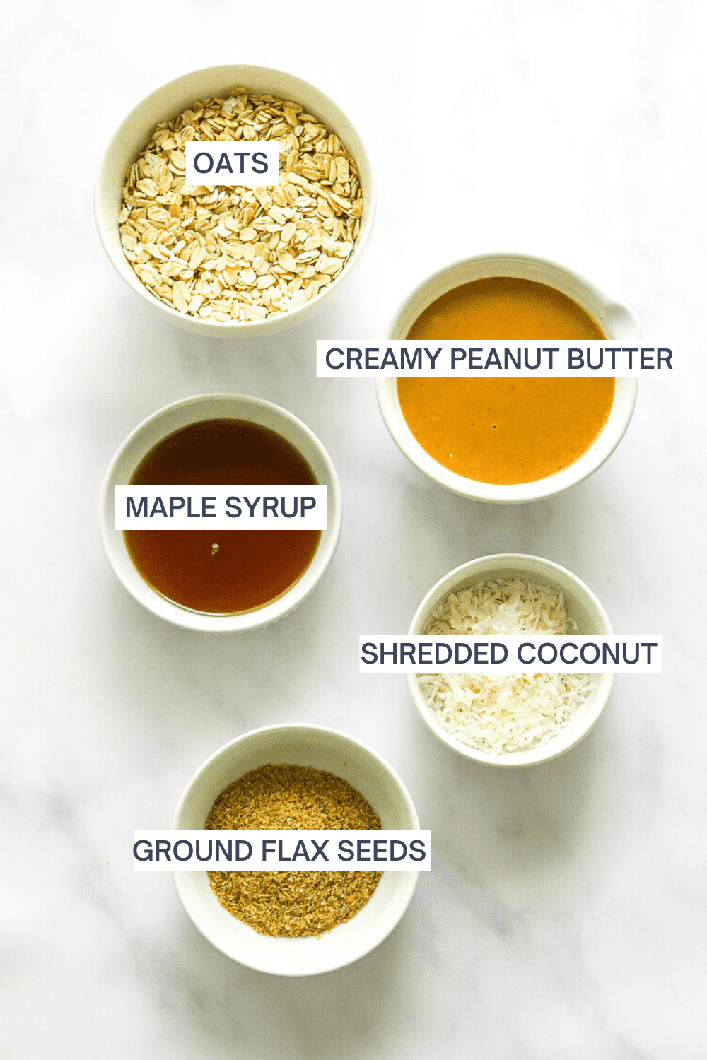Oats in a bowl with a bowl of creamy peanut butter, a bowl of ground flax seeds, shredded coconut and a bowl of maple syrup in front of it with labels over each ingredient. 