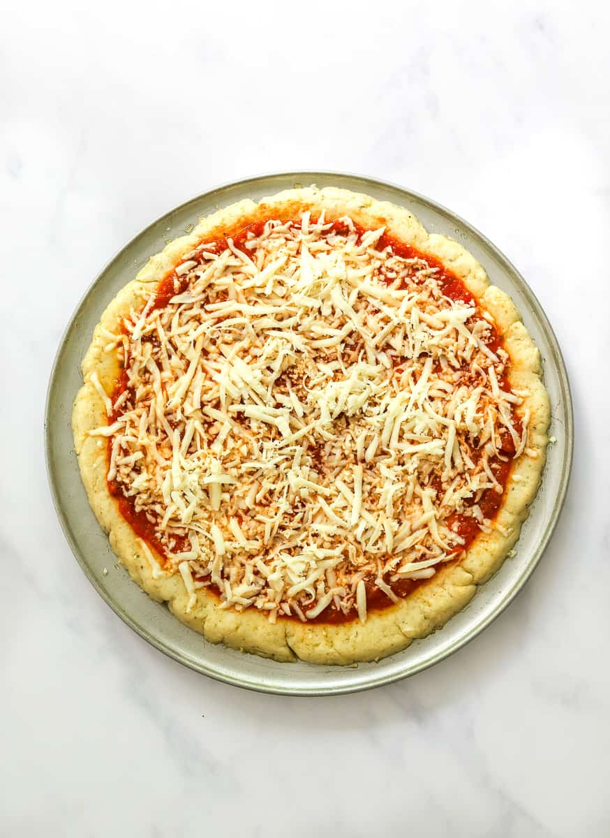 Pizza crust topped with sauce and unmelted cheese on a round pizza pan.