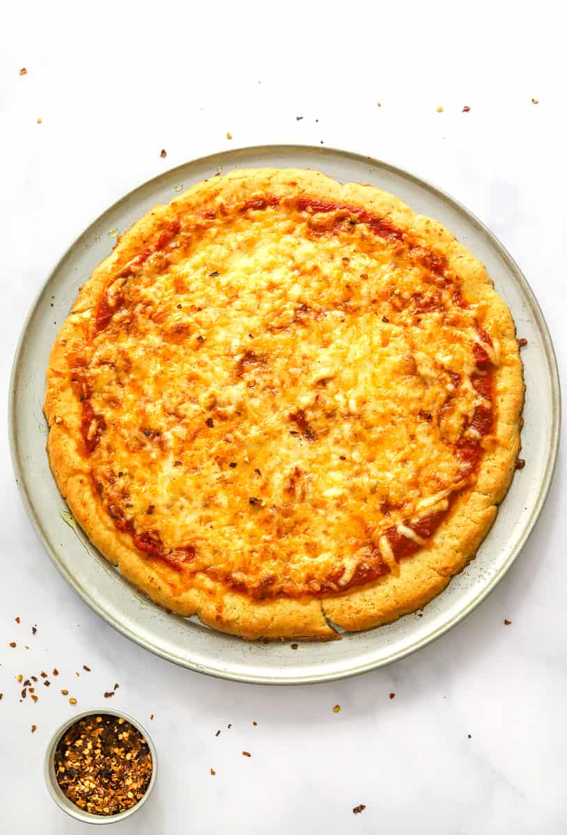 Cooked cheese pizza on a round pizza pan with a small bowl of red pepper in front of it.