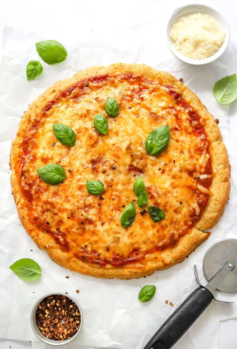 Cooked protein pizza topped with basil leaves with a bowl of grated parmesan cheese behind it and a bowl of red pepper flakes in front of it.