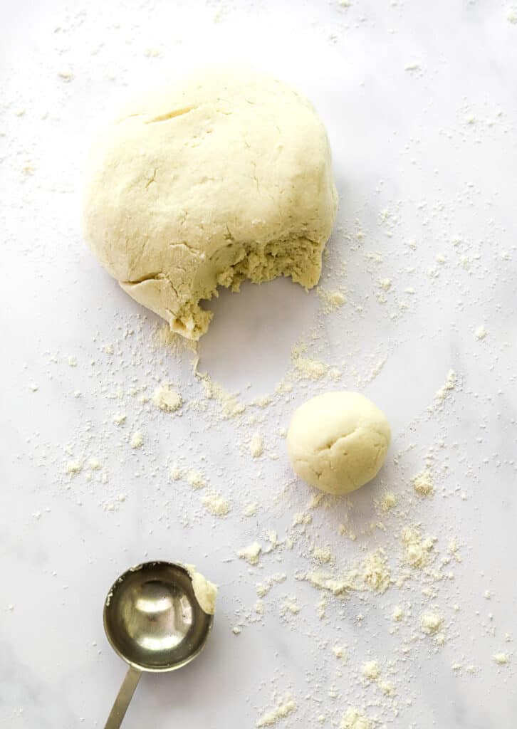 Dough ball with some pulled off of it rolled into a ball next to it with some flour scattered around it and a round metal measuring spoon in front of it.