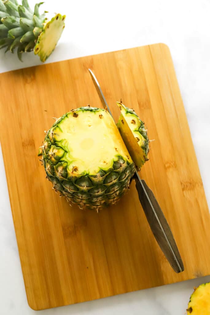 Pineapple on a brown cutting board with the top cut of and a knife slicing the skin off of it.