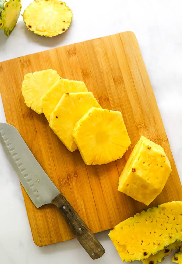 Slices of fresh pineapple on a brown cutting board with a kitchen knife on the board next to it.