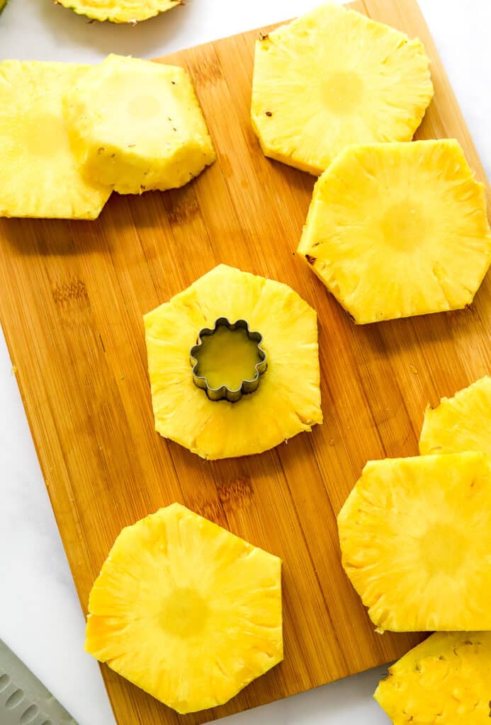 Slices of pineapple on a brown cutting board with one in the center with a small cookie cutter cutting the core of the pineapple slice out of it.