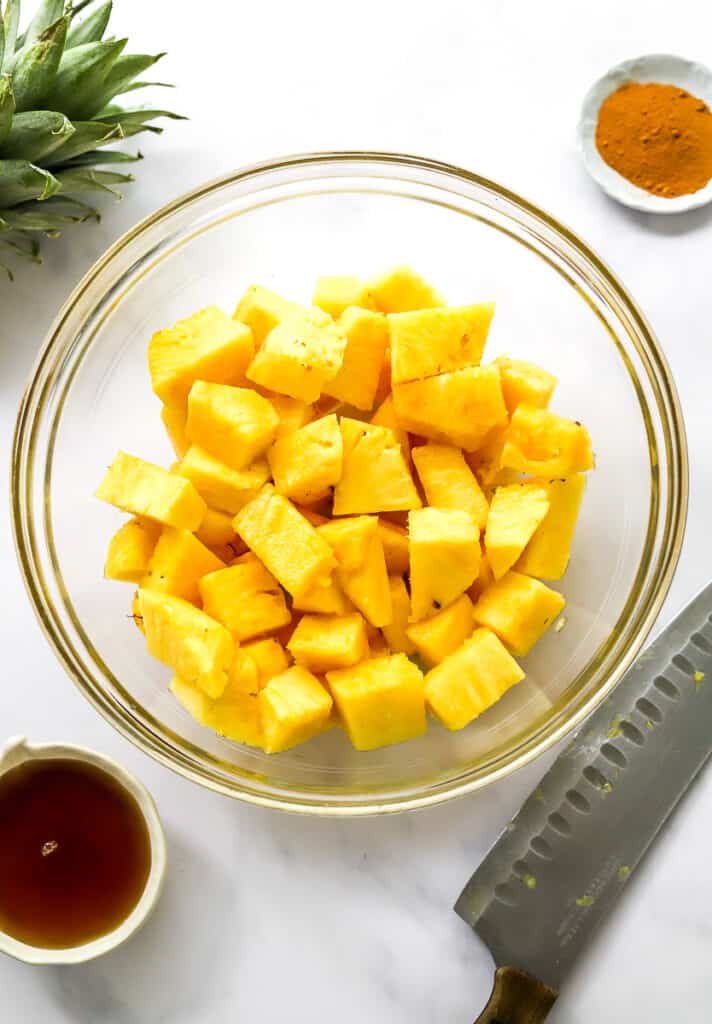 Cubed pineapple chunks in a glass bowl with a knife and bowl of syrup in front of it and a bowl of ground cinnamon and the top of the pineapple behind it.