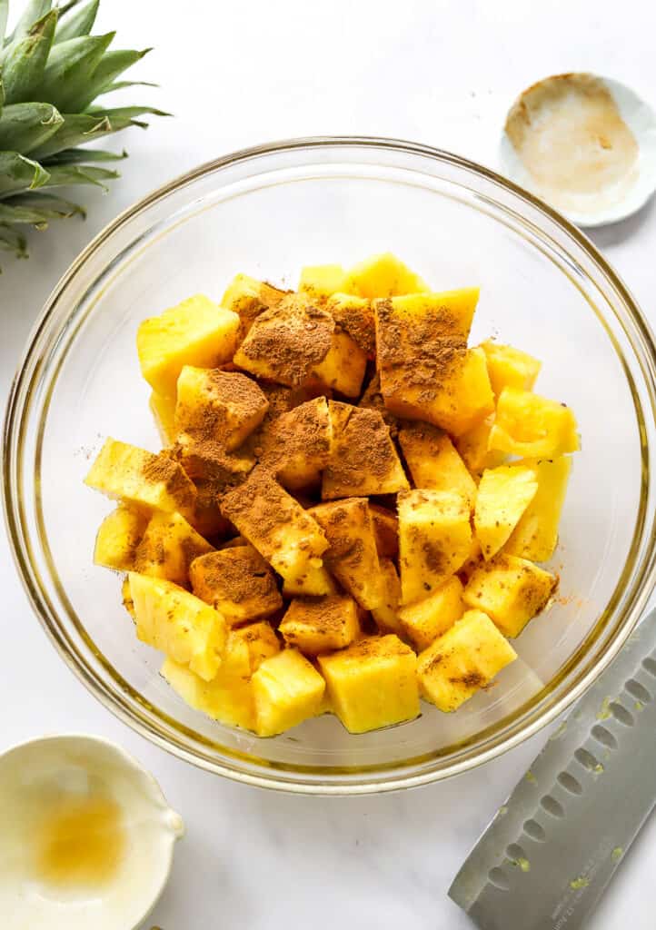 Bowl of cubed pineapple with maple syrup and ground cinnamon on it with a knife and empty bowl in front of ti and a white small bowl and head of pineapple behind it.