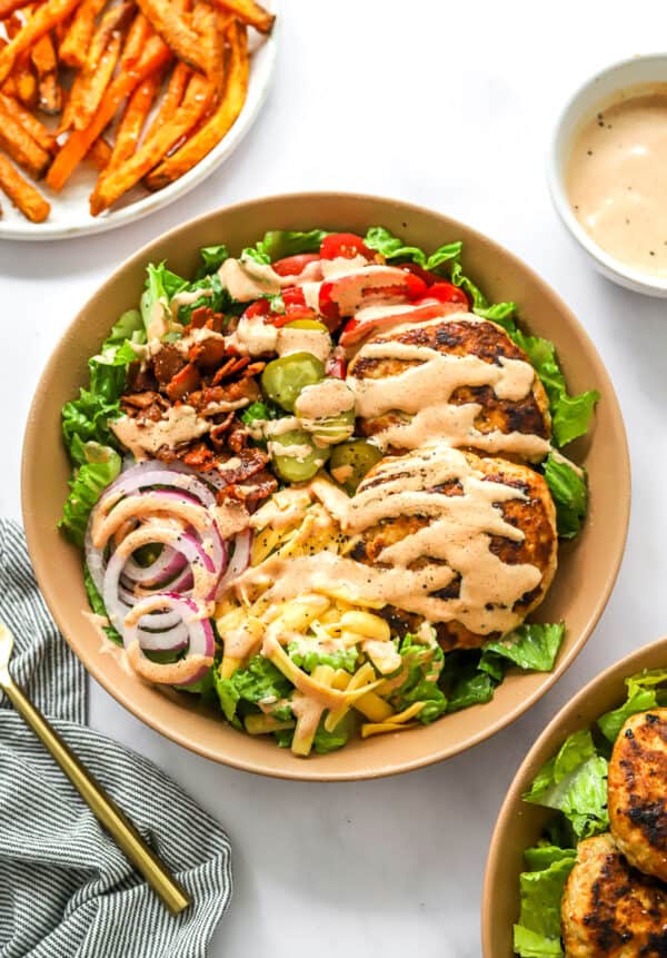 Finished turkey burger salad bowl drizzled with burger sauce with another bowl of it in front of it and a plate of fries and more sauce in a bowl behind it.