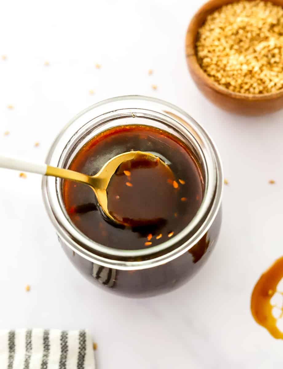 Glass jar of brown sticky sauce with a spoon in the jar and sesame seeds in a bowl behind it.