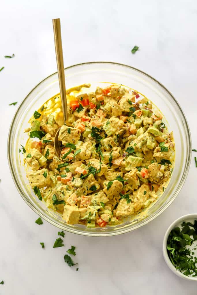 Mixed chicken salad with creamy dressing and herbs in a glass bowl with a spoon in it and a bowl of herbs in front of it.