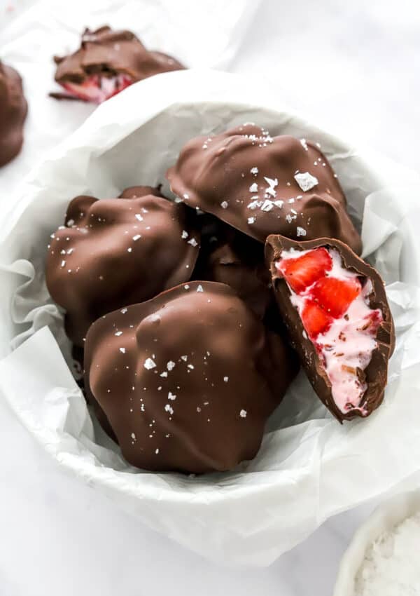 Chocolate bites with yogurt and strawberries in them in a bowl on top of white paper with more clusters behind it.
