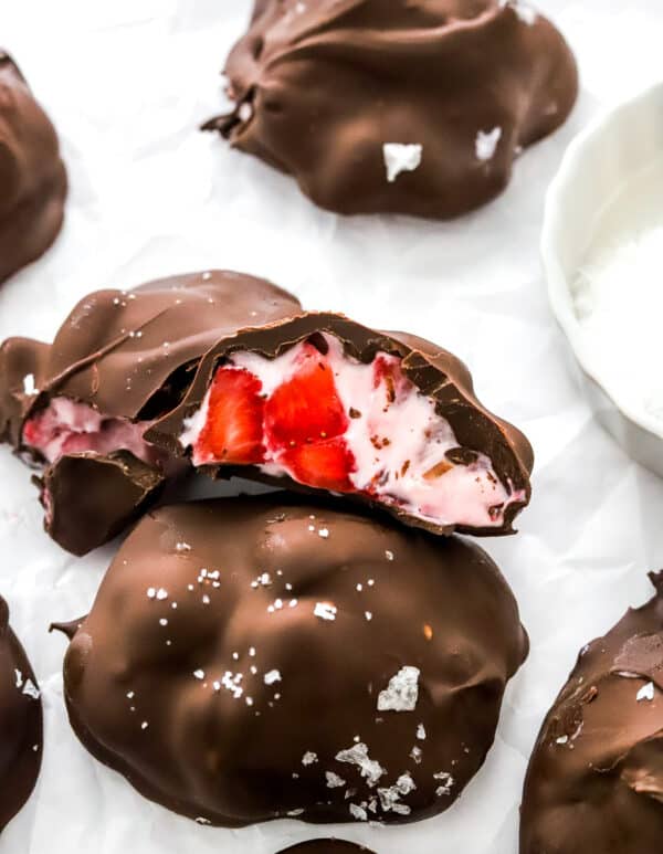 Strawberry yogurt filled chocolate treats with one cut open on white paper with a white bowl of seas salt next to them.