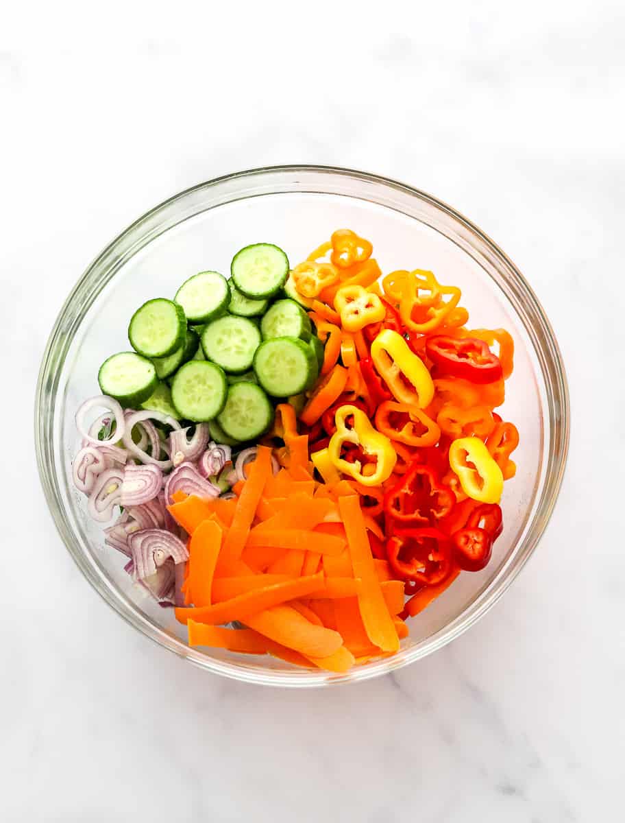 Glass bowl filled with sliced cucumber, bell pepper, shallots and shaved carrots.
