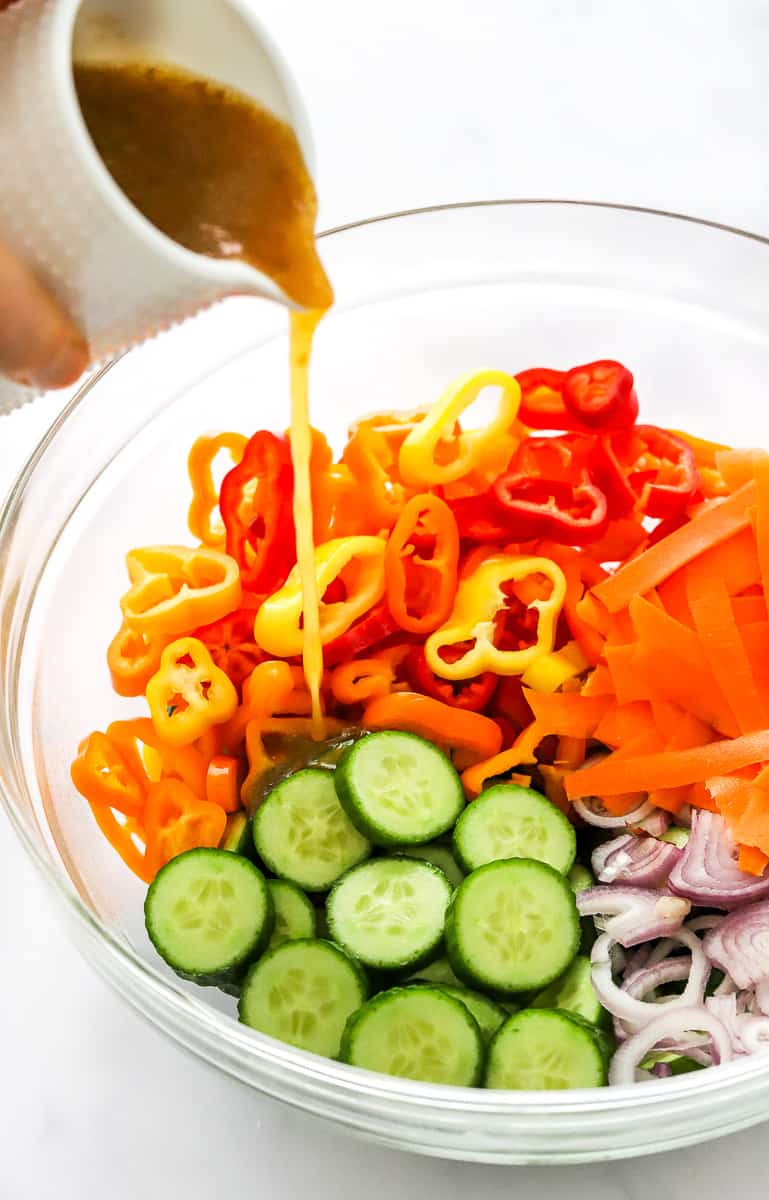 Salad dressing being pored into a glass mixing bowl filled with cucumber, pepper rings, carrots and onion.