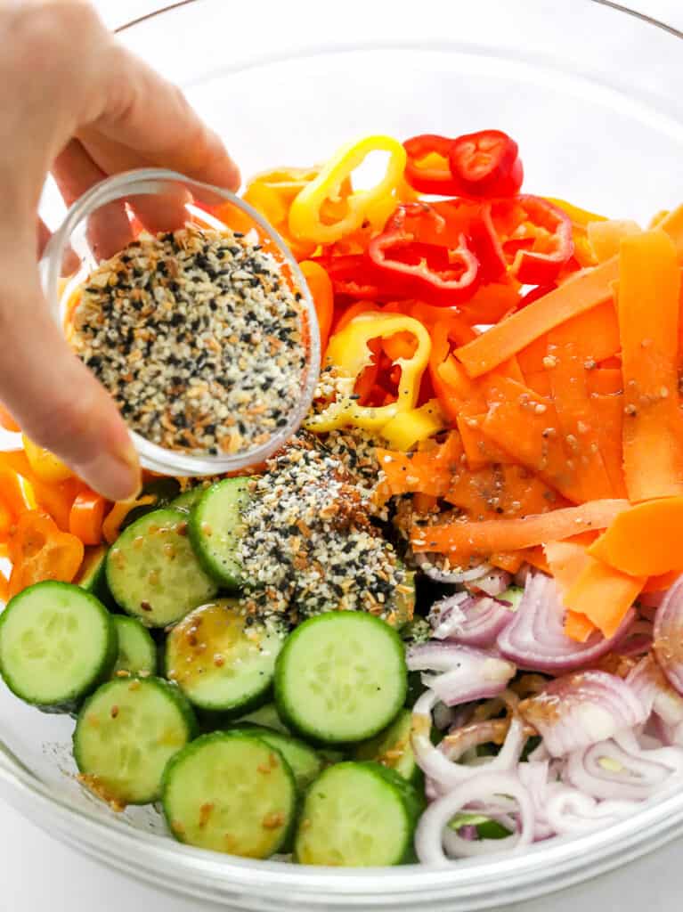 Everything bagel seasoning is sprinkled onto cucumber, peppers, onion and carrots, and chopped in a glass bowl.