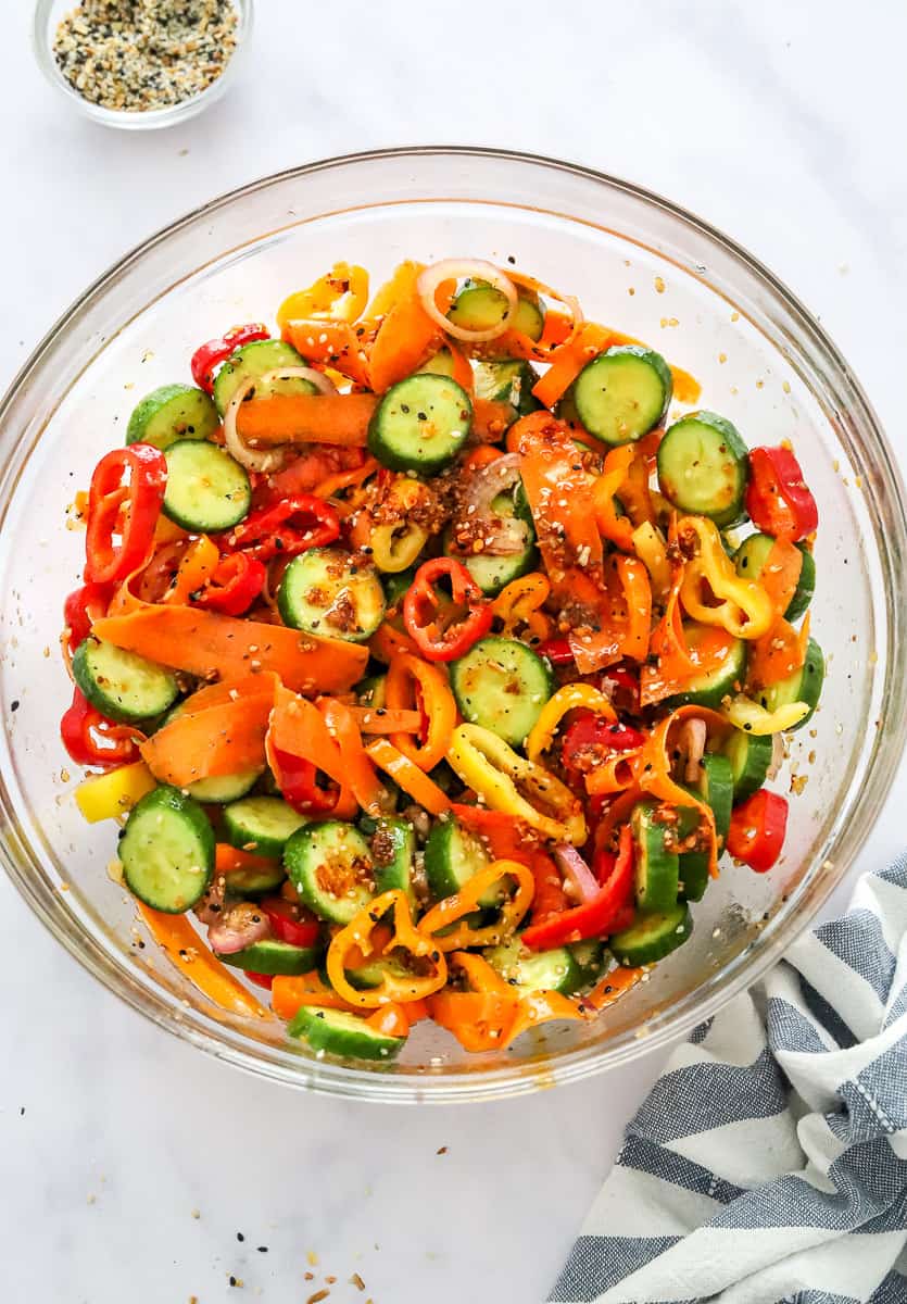 Glass mixing bowl filled with mixed up pepper, cucumber salad with a bowl of seasoning behind it, and striped towel in front of it.