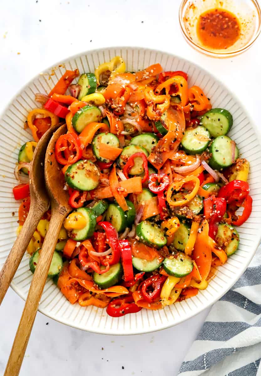 Sliced Persian cucumbers, sliced baby peppers, onion, and carrots mixed with dressing in a beige salad bowl with wood spoons in the bowl with a bowl of oil behind it and blue striped linen in front of it.