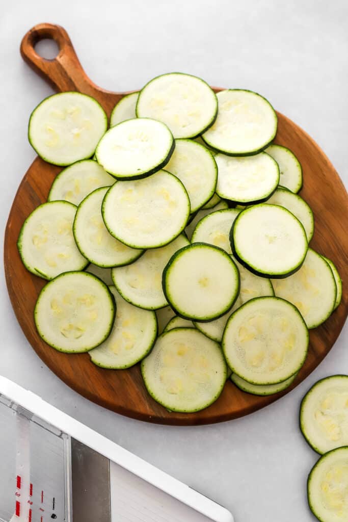 Sliced zucchini on a round wood cutting board with a mandolin in front of it.