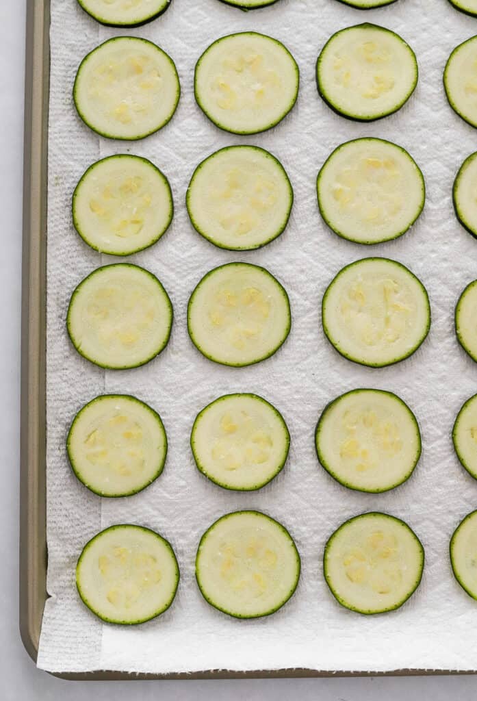 Sliced zucchini slices in rows on top of a towel paper-lined baking sheet.