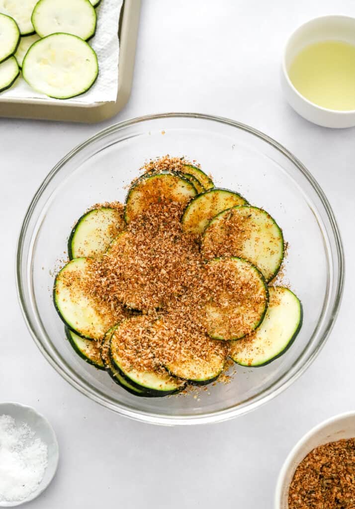 Raw zucchini sliced in a glass bowl with parmesan seasoning on it with more zucchini slices and oil behind it and a bowl of salt and more parmesan seasoning in front of it.