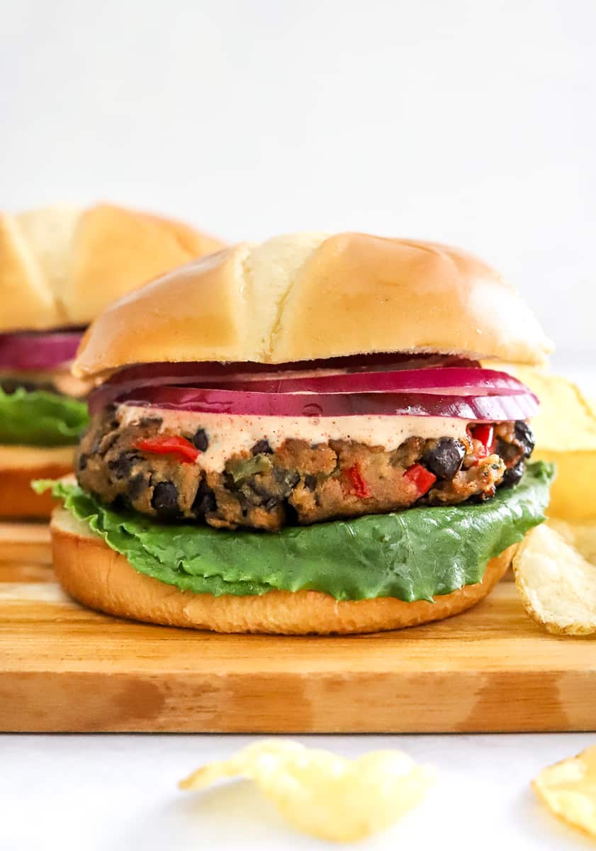 Black bean veggie burgers piled inside of a white bun with lettuce and tomato on a cutting board with a few chips in front of it.