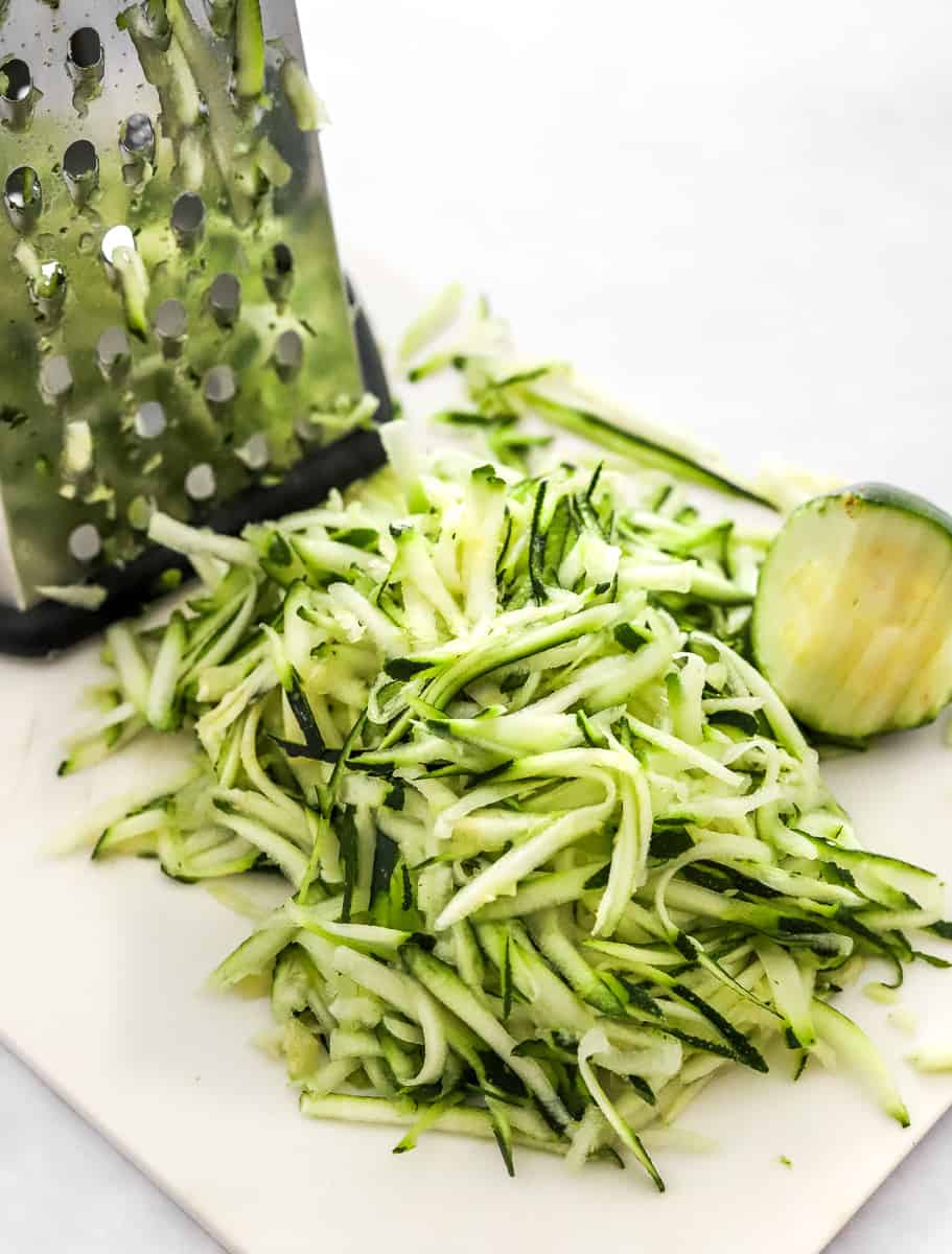 Shredded raw zucchini on a white board with a silver grater next to it.