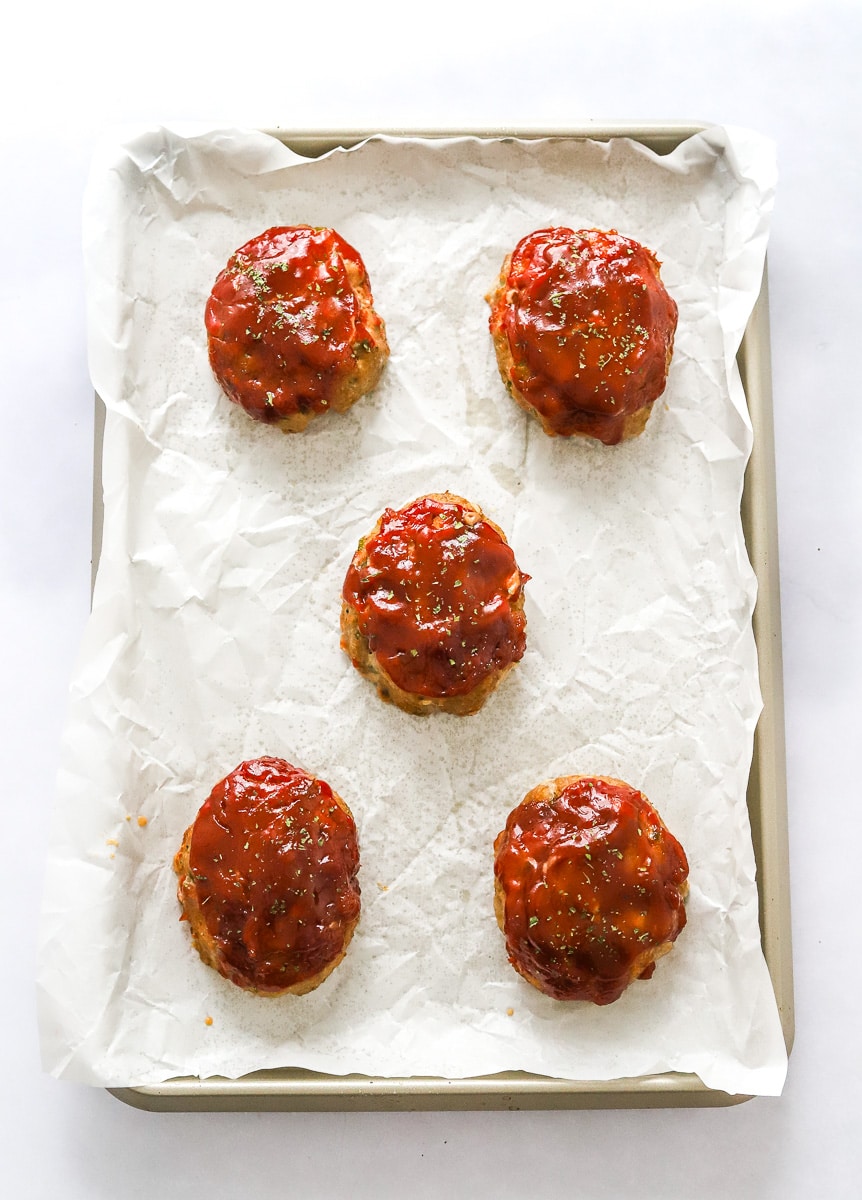 5 cooked individual meatloaves toped with tomato glaze on a parchment paper lined baking sheet.